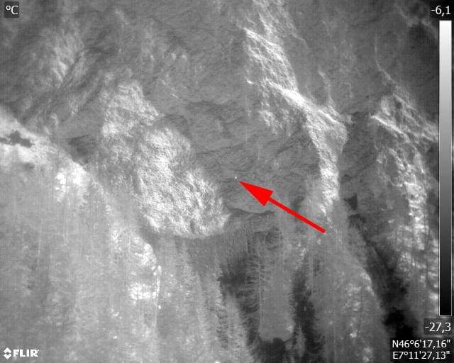 Thermal camera frame with a red arrow marking a faint heat signature on cliffs.