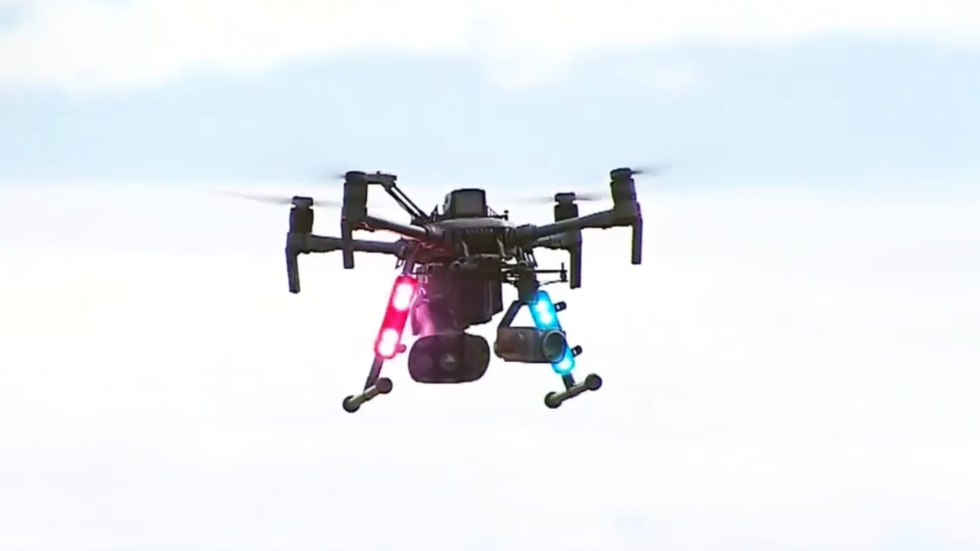 A police drone with a loudspeaker and colored strobes hovers in an overcast sky.