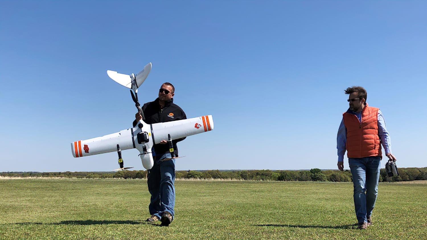 Two people carry a fixed-wing test drone across a grassy field.