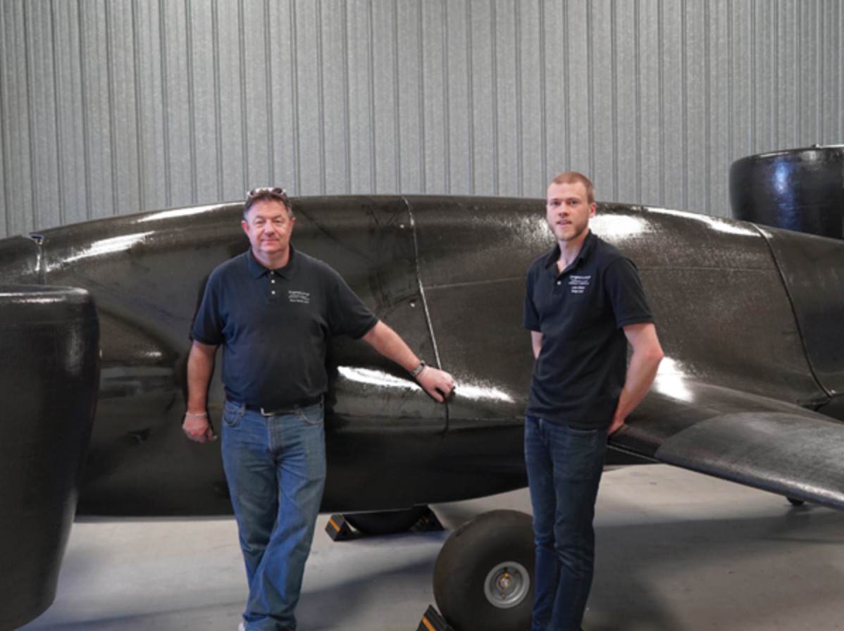 Two Sabrewing team members standing beside the black Rhaegal prototype in a hangar.