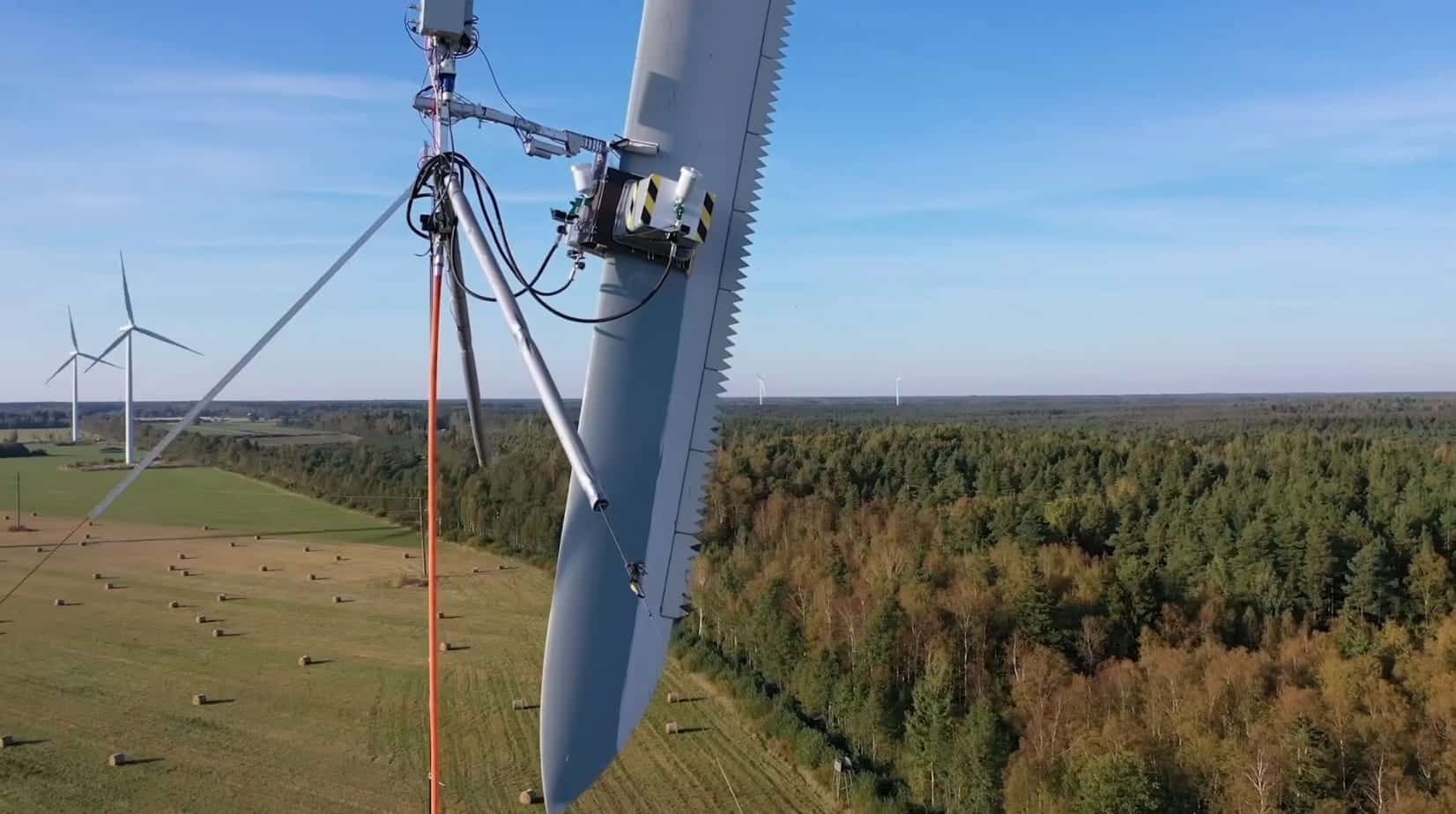 Robotic maintenance rig climbing a wind turbine blade above farmland and forest.