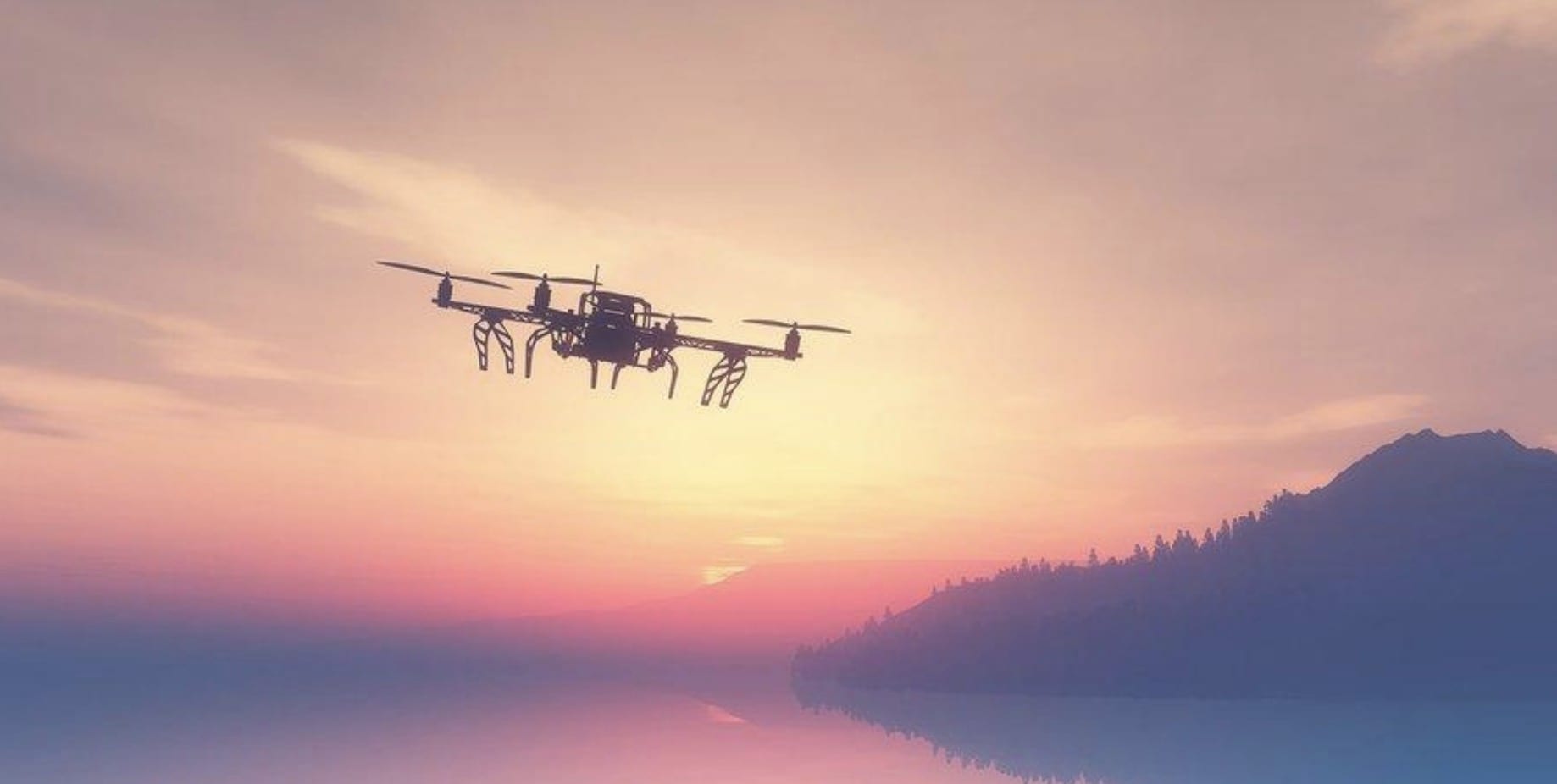 A hexacopter silhouetted against an orange sunset above calm water and hills.