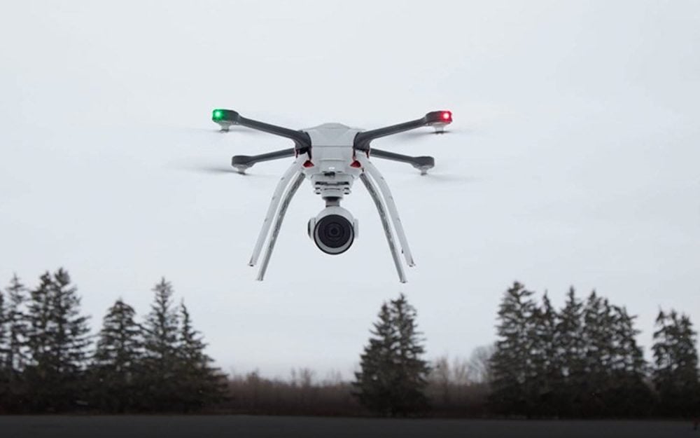 A white quadcopter with navigation lights hovering above a treeline under grey sky.