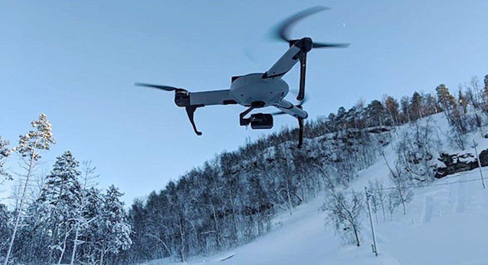 A white quadcopter hovers over a snow-covered hillside in winter light.