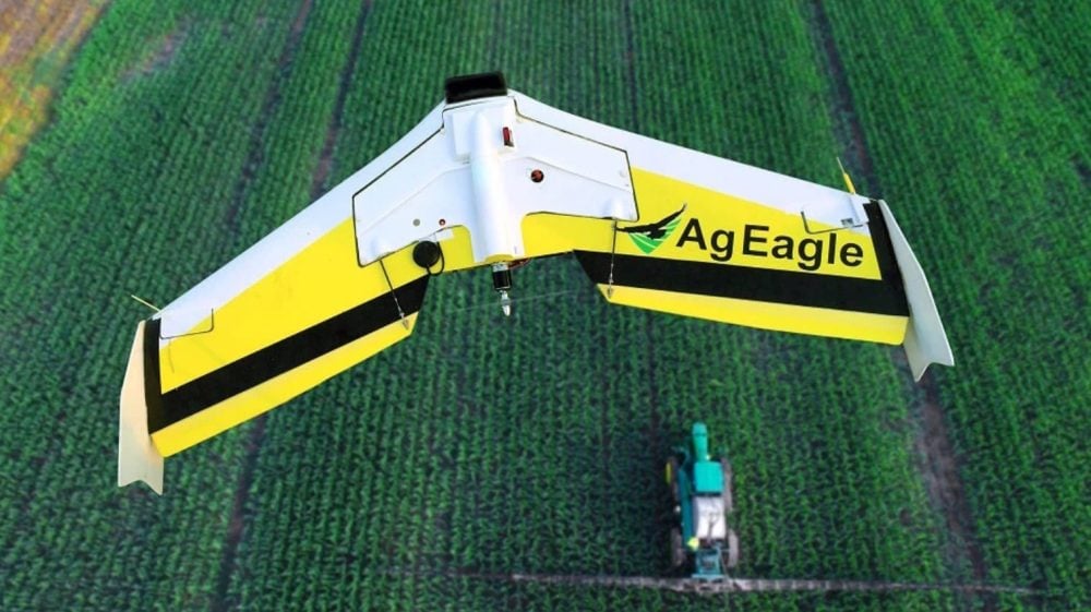 An AgEagle fixed-wing survey drone banking over a green crop field as a sprayer works below.