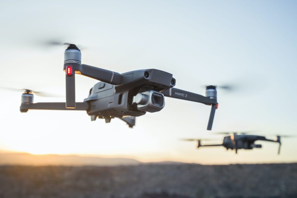 Two DJI Mavic 2 drones hovering against a desert sunset, one sharp and one behind.