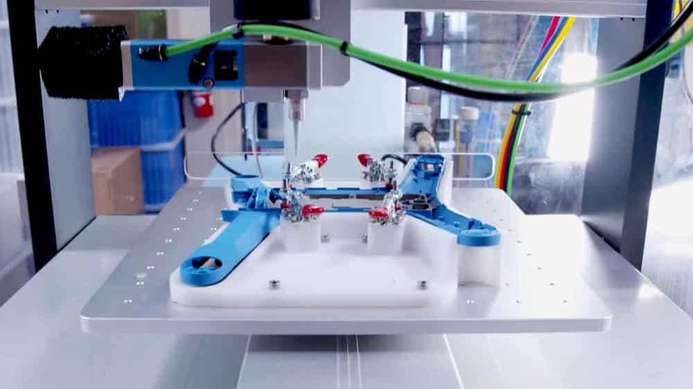 Robotic arm assembling a blue drone airframe on a jig inside a production cell.