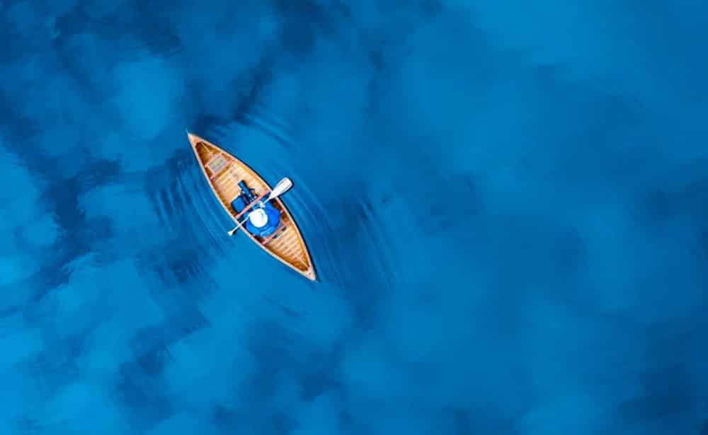 Overhead aerial of a lone paddler in a wooden canoe on deep blue water.