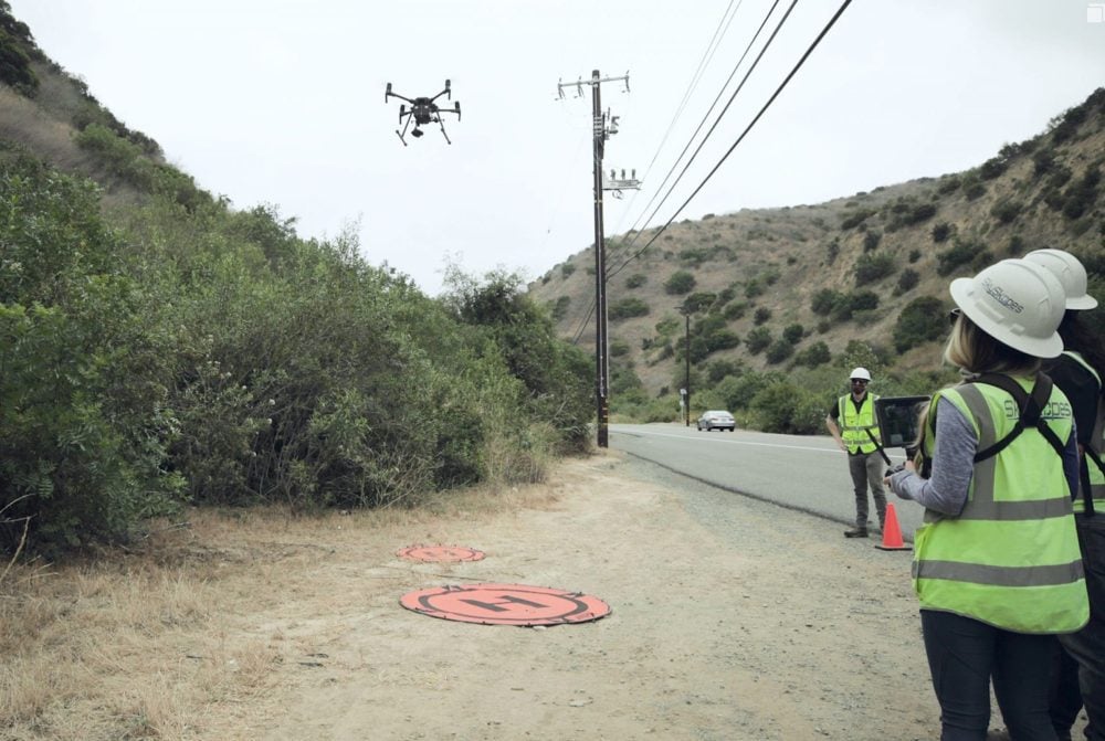 Utility crew in high-visibility vests fly a quadcopter beside hillside power lines along a coastal road.