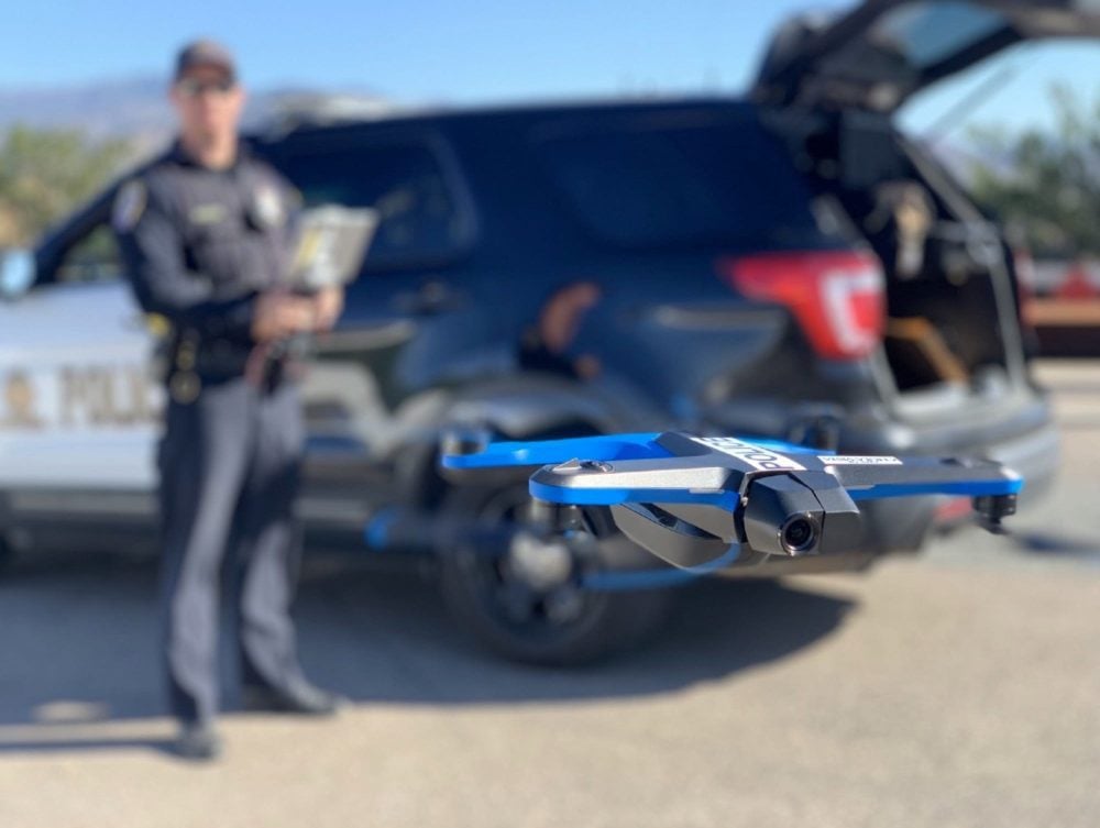 A blue Skydio 2 hovers in front of a police SUV as an officer watches.