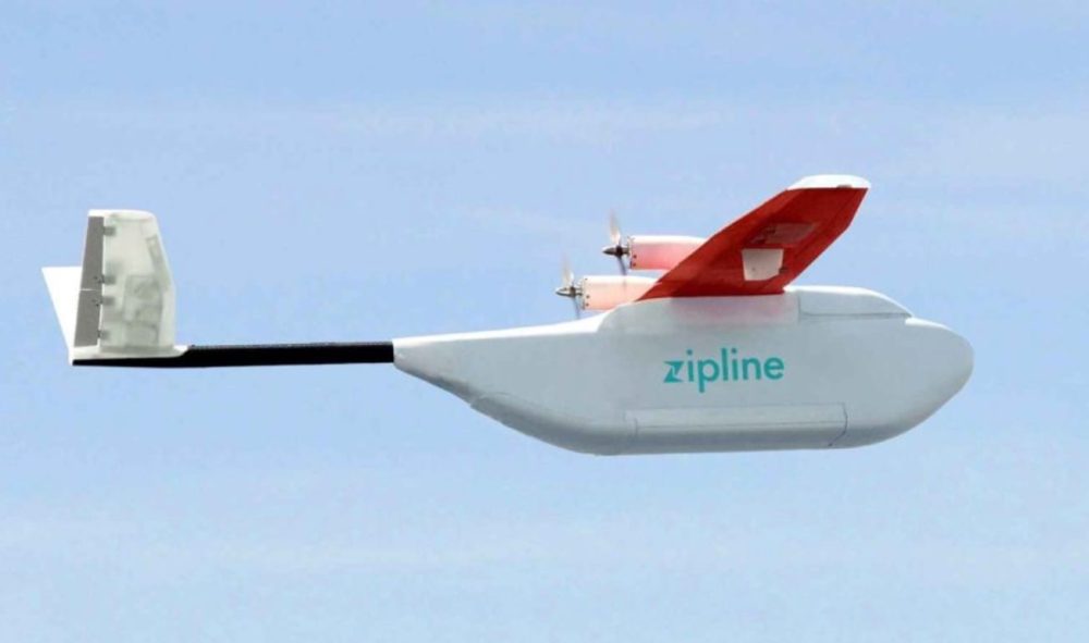 A white Zipline fixed-wing delivery drone with a red tail cruising against clear blue sky.