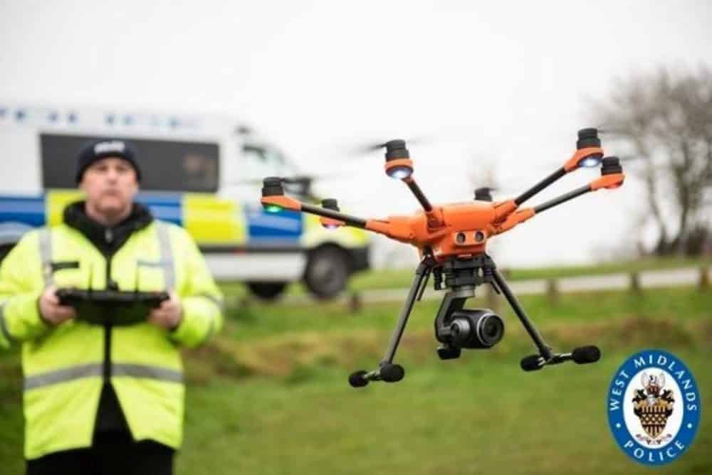 West Midlands Police officer in a hi-vis jacket flying an orange hexacopter beside a police van.