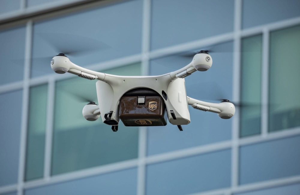 White Matternet quadcopter carries a UPS package box past a glass office building.