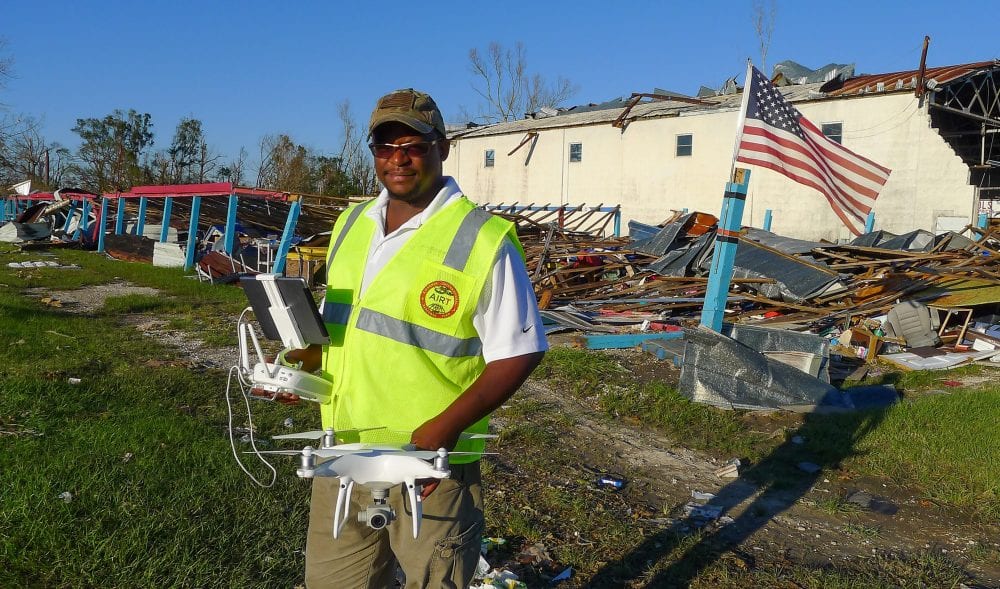 AIRT volunteer in a safety vest holds a Phantom drone amid hurricane wreckage.