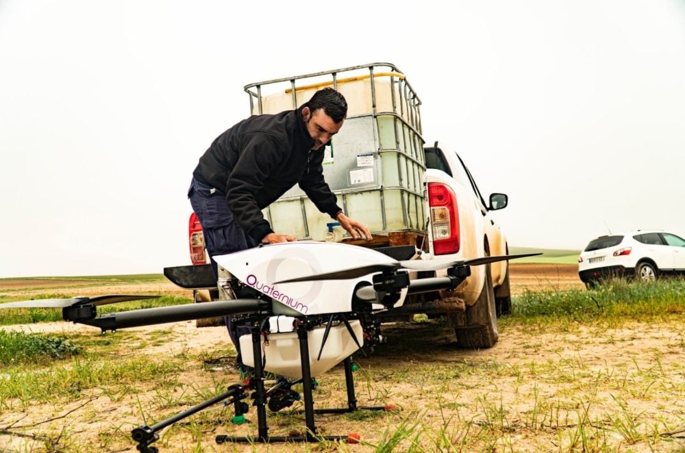 Technician refills a large white Quaternium spraying drone from a tank on a pickup.