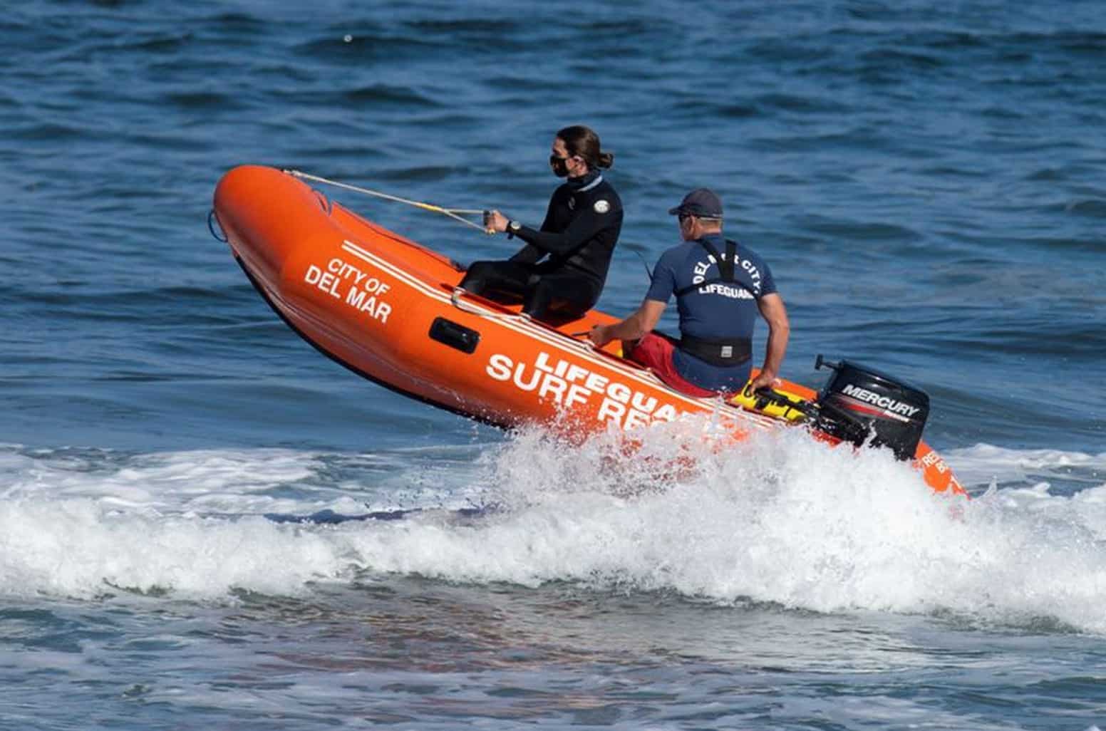 Two City of Del Mar lifeguards riding an orange surf rescue inflatable through the waves.