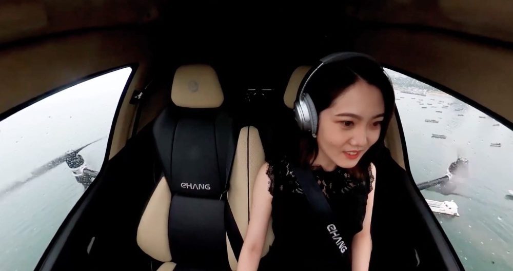 Passenger wearing headphones sits inside an EHang 216 cabin as boats and coastline pass below.
