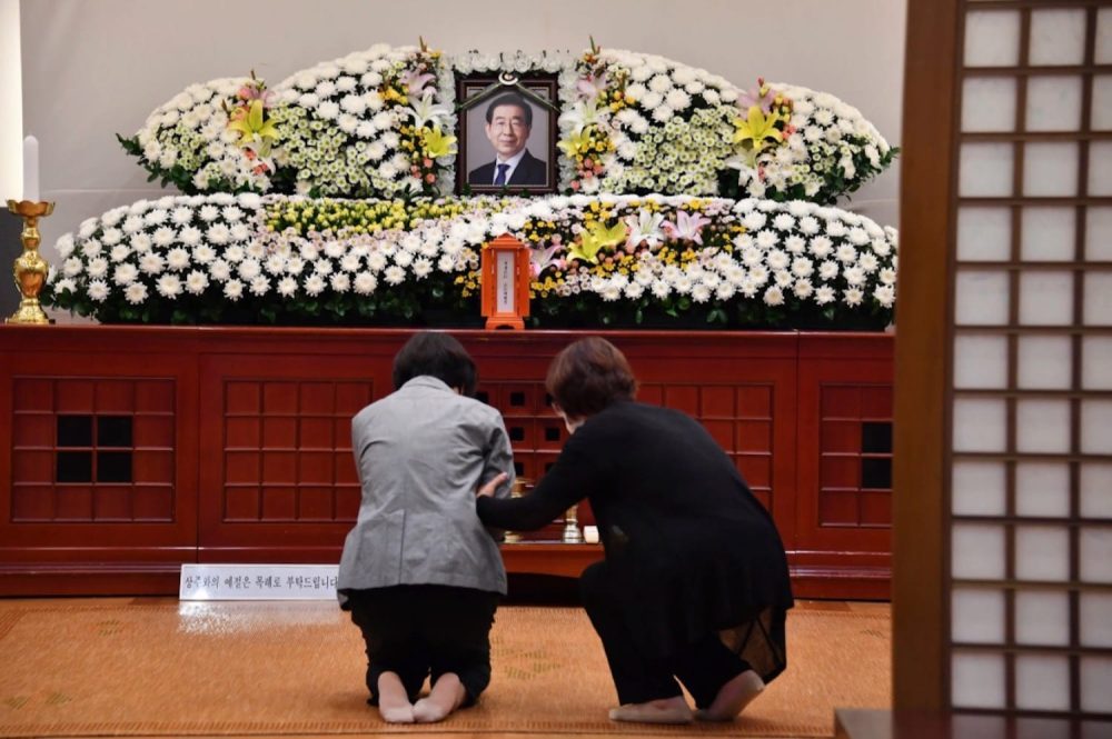 Two mourners kneel before a flower-covered memorial altar holding a portrait of Seoul Mayor Park Won-soon.