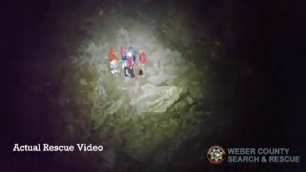 Night aerial view of Weber County rescuers gathered around a person under a searchlight.