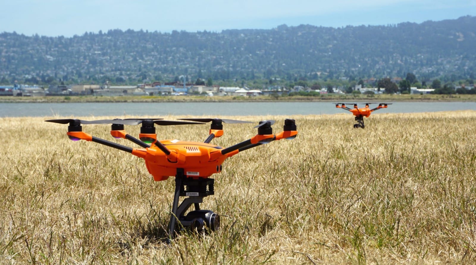 Two orange hexacopters with gimbal cameras sitting in dry grass beside a bay.