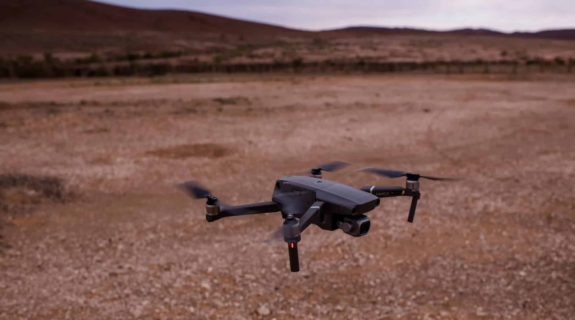 DJI Mavic 2 hovers low over dry red outback scrub under an overcast sky.