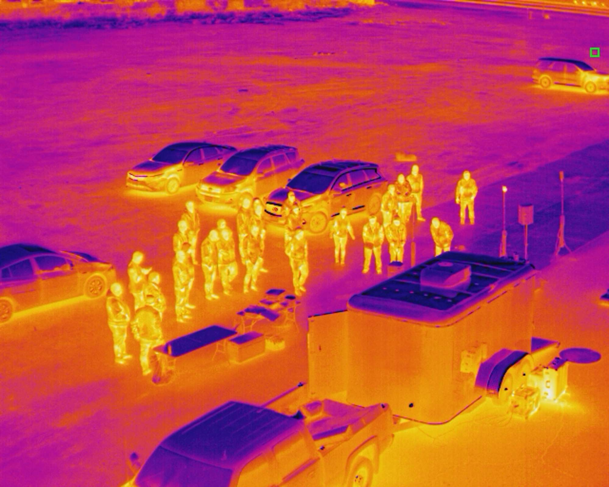 Thermal aerial view of people standing among parked vehicles and equipment cases.