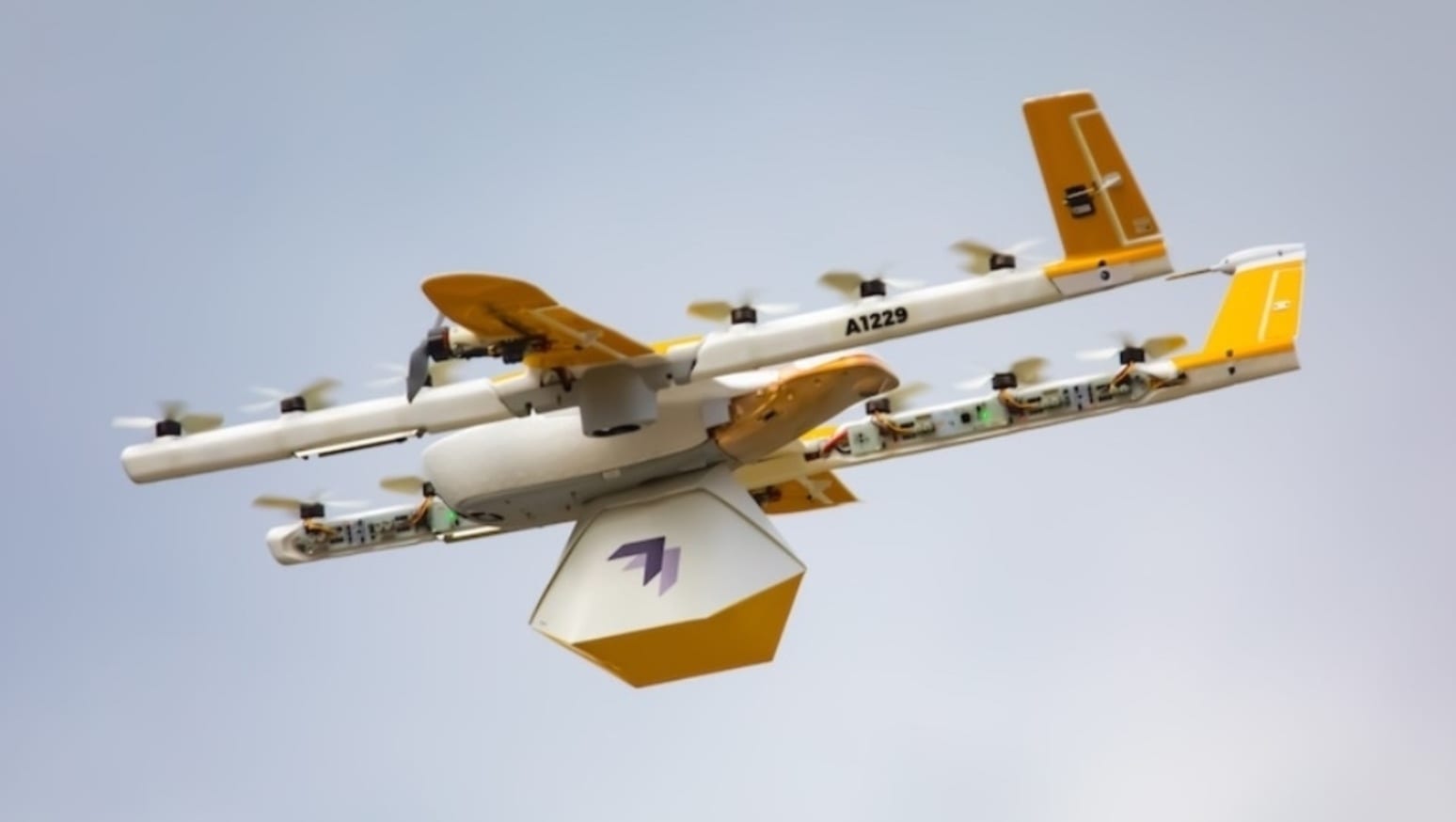 Wing delivery aircraft in flight carrying a package beneath its yellow fuselage.