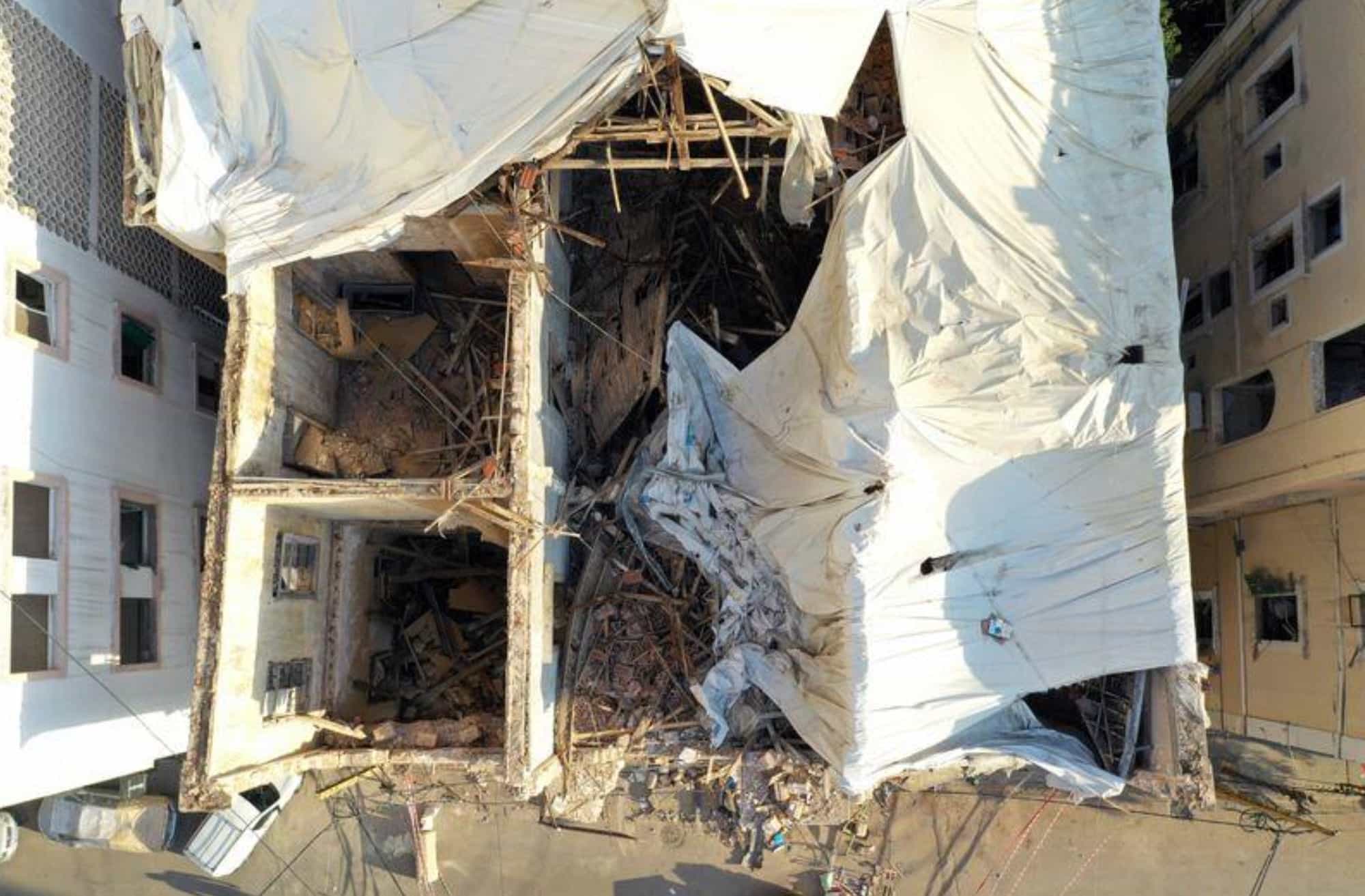 Overhead drone view of a gutted building draped in torn white tarpaulins over rubble.
