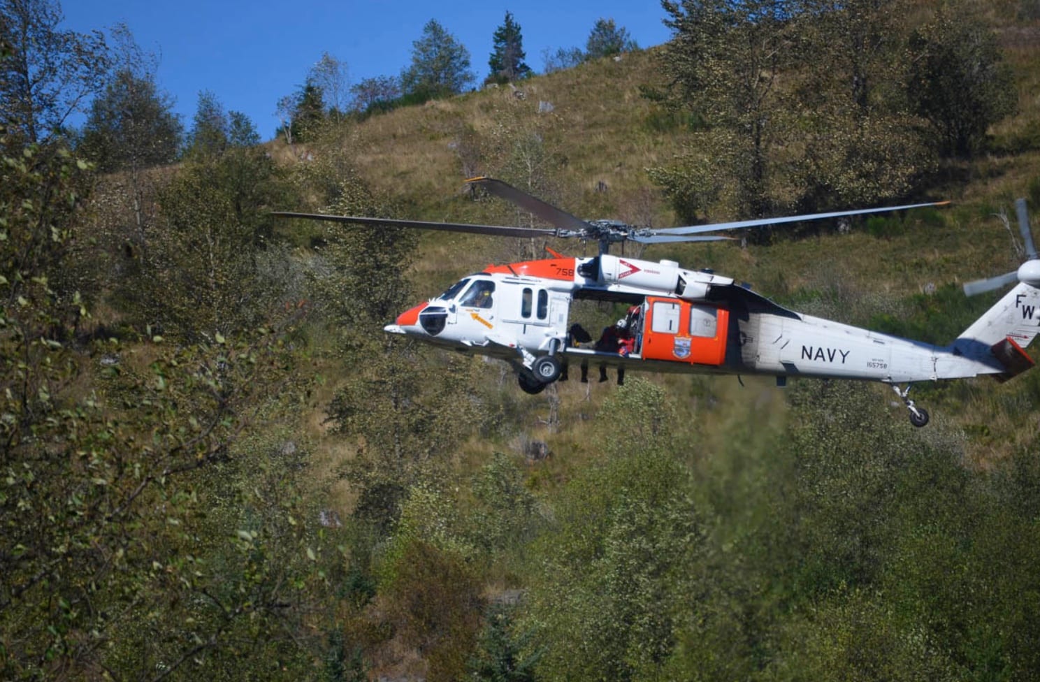 Navy search helicopter flying low over forested slopes.
