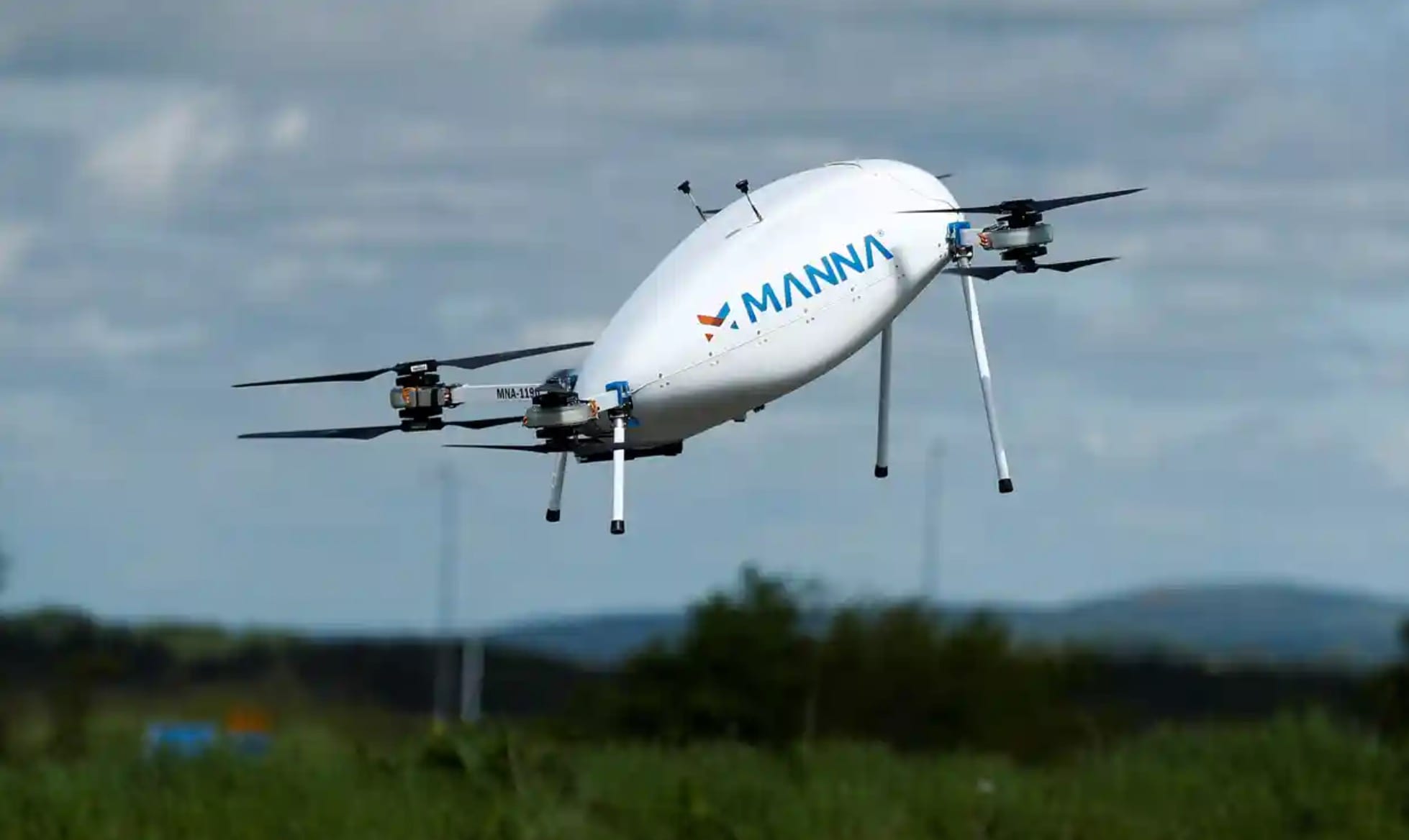 Manna Aero delivery drone hovers above green fields under a cloudy sky.