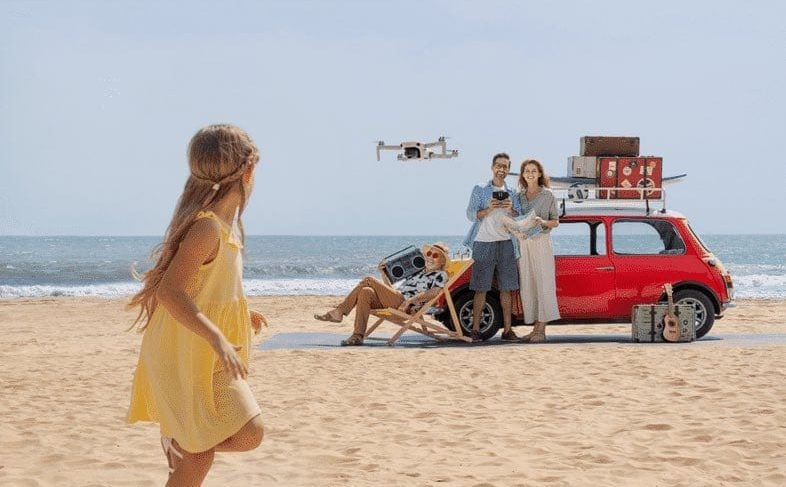 Family and a red car on a beach as a mini drone hovers.