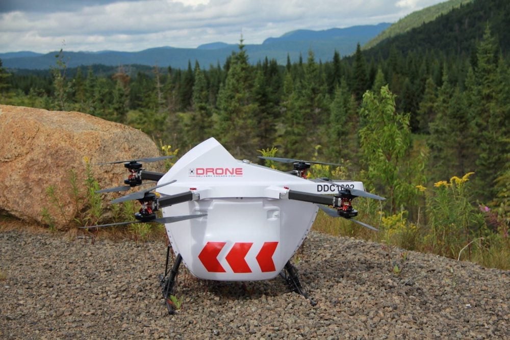 A white Drone Delivery Canada cargo drone parked on gravel before forested mountains.