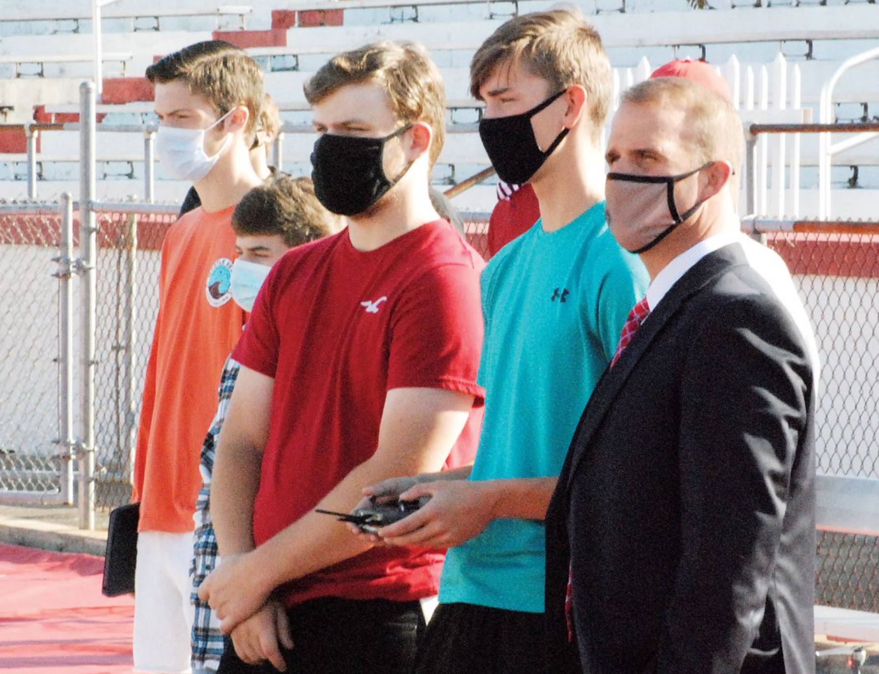 Masked high school students line up trackside while one holds a drone controller.