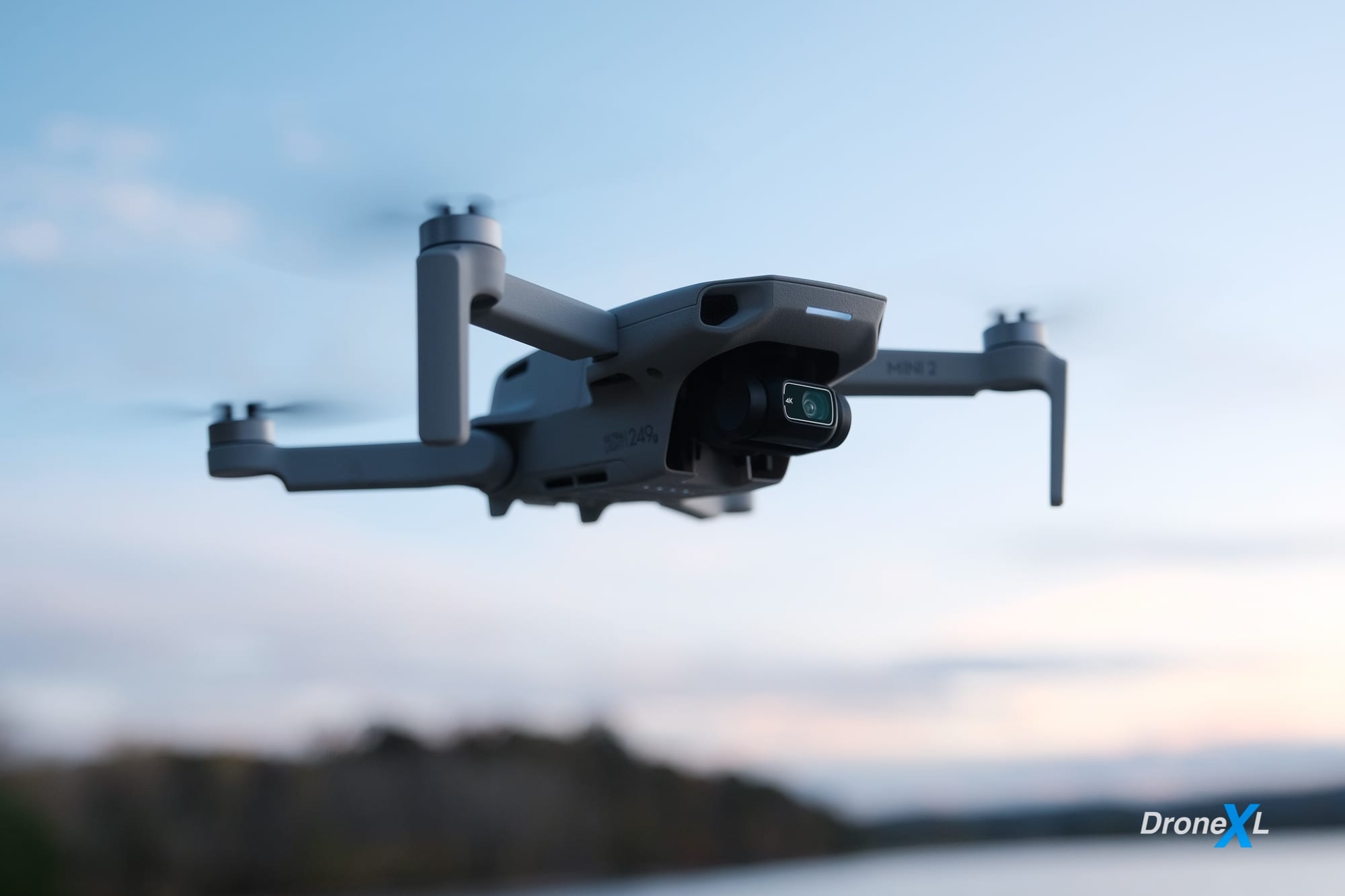 A DJI Mini 2 hovering in blue sky above a distant lakeshore.