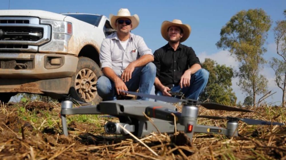 Two ranchers in cowboy hats crouching beside a pickup truck and a Mavic drone.