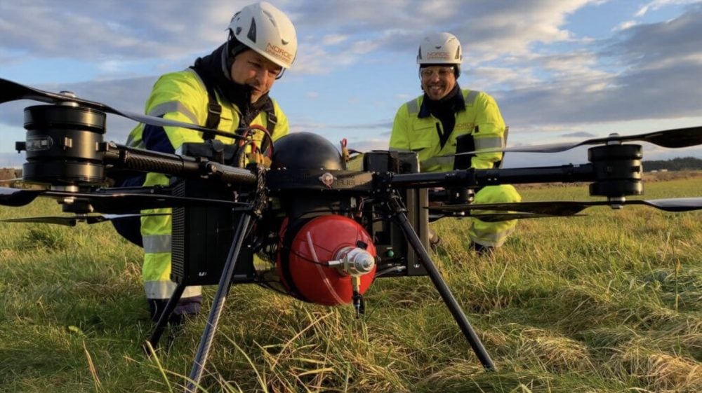 Two technicians in high-visibility gear prepare a large multirotor carrying a red hydrogen tank.
