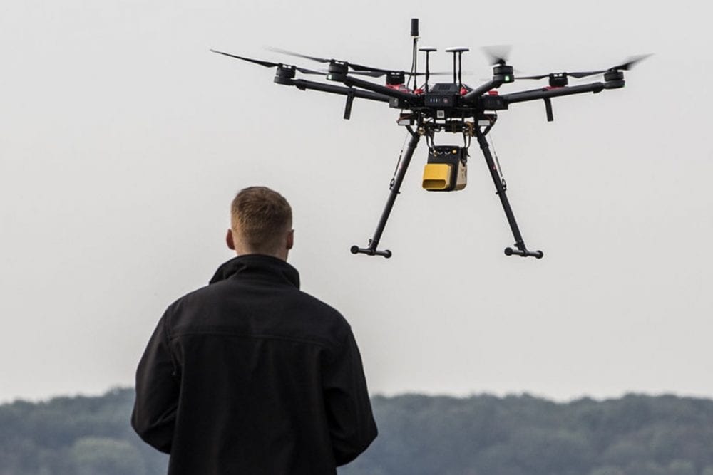 A pilot watches a black hexacopter carrying a yellow payload hover overhead.