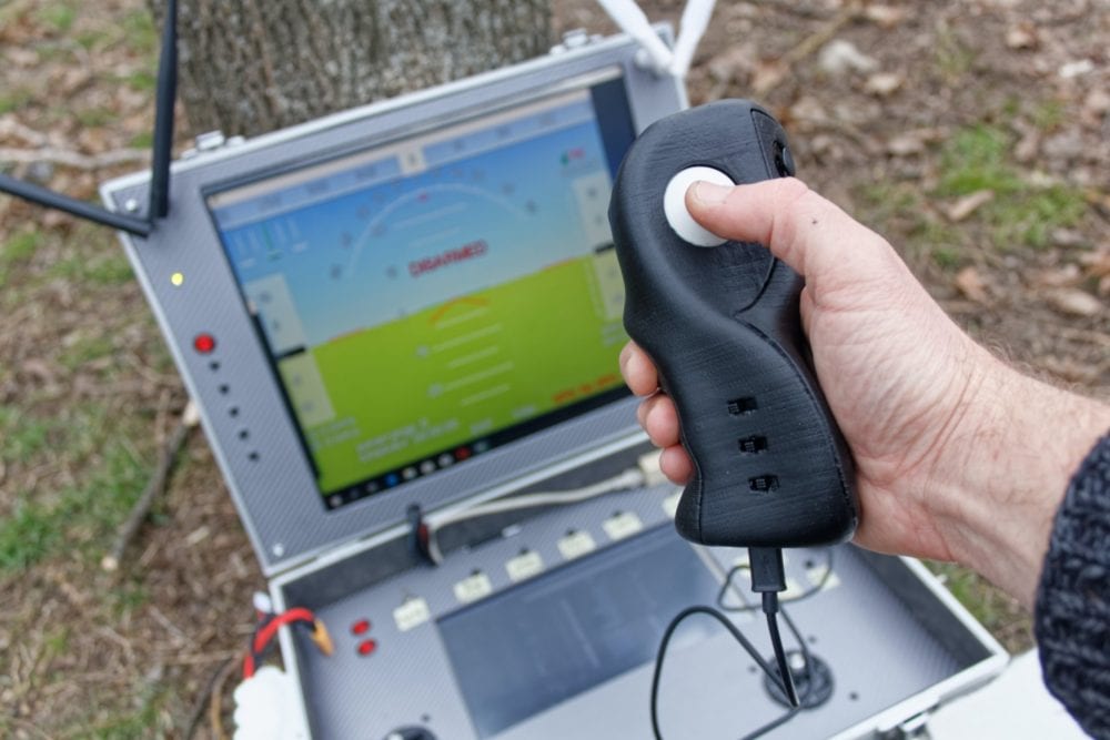 Hand gripping the one-handed Knuckles-5 controller beside a ground station screen.