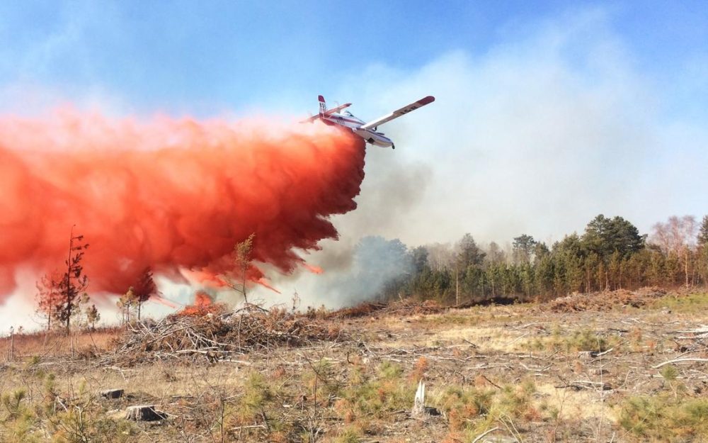 An air tanker drops red retardant over a smoking forest near cleared ground.