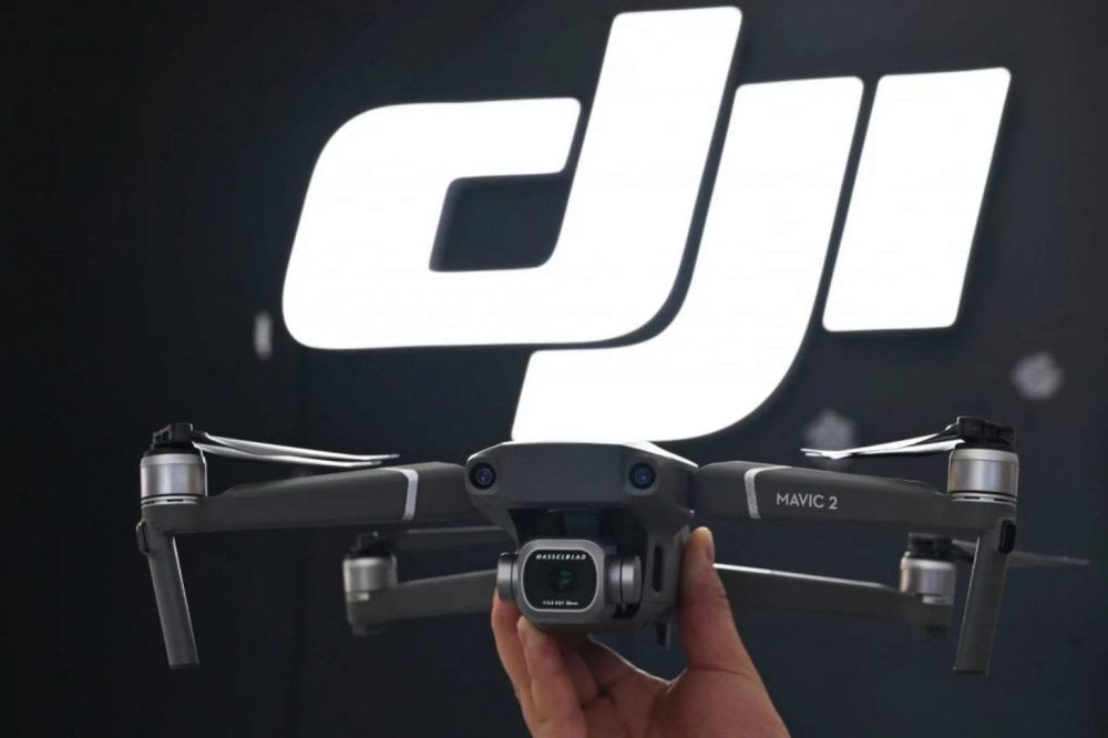 Hand holds a DJI Mavic 2 in front of an illuminated DJI logo wall.
