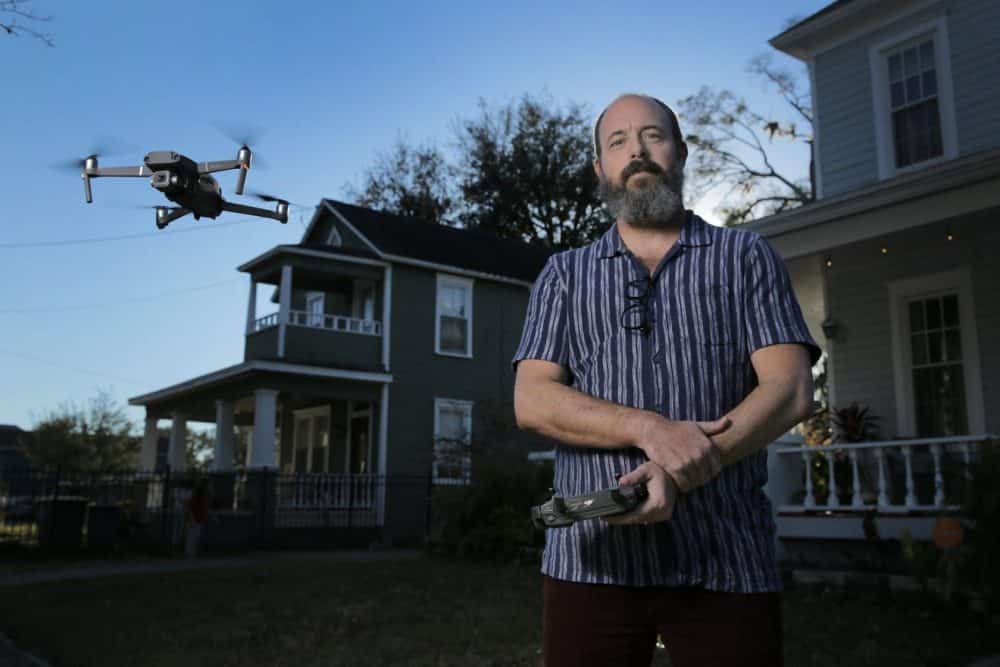 Aerial photographer holds a controller while his Mavic hovers outside a home.