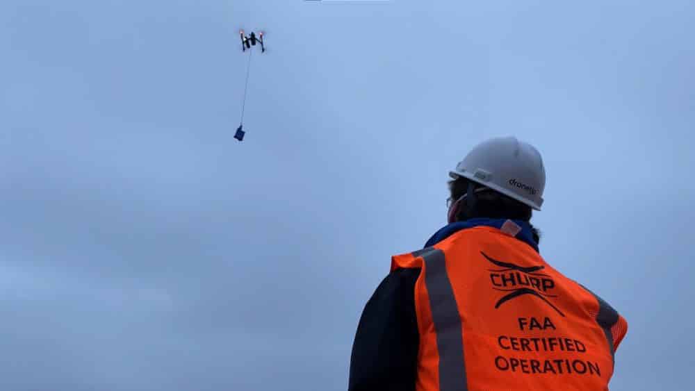 A hard-hatted operator watches a delivery drone lower a package on a tether overhead.