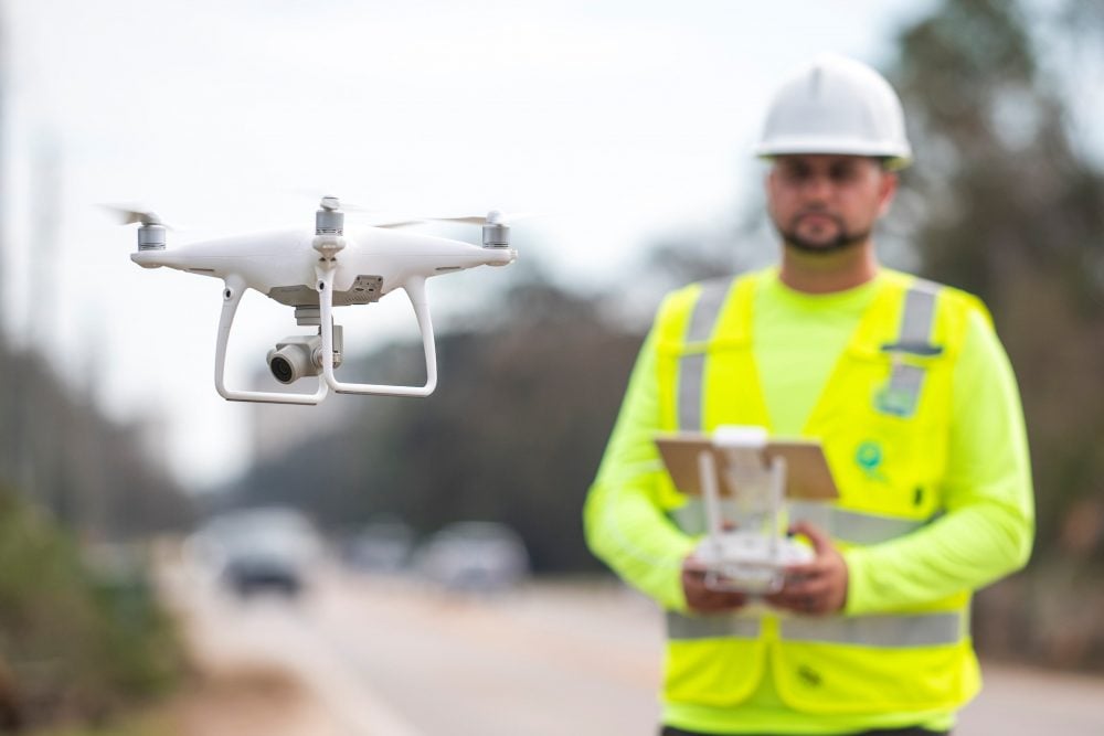 A worker in a hard hat and hi-vis vest flies a DJI Phantom roadside.