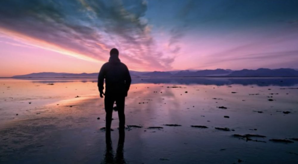 Silhouetted figure standing on a mirror-like salt flat at sunset.