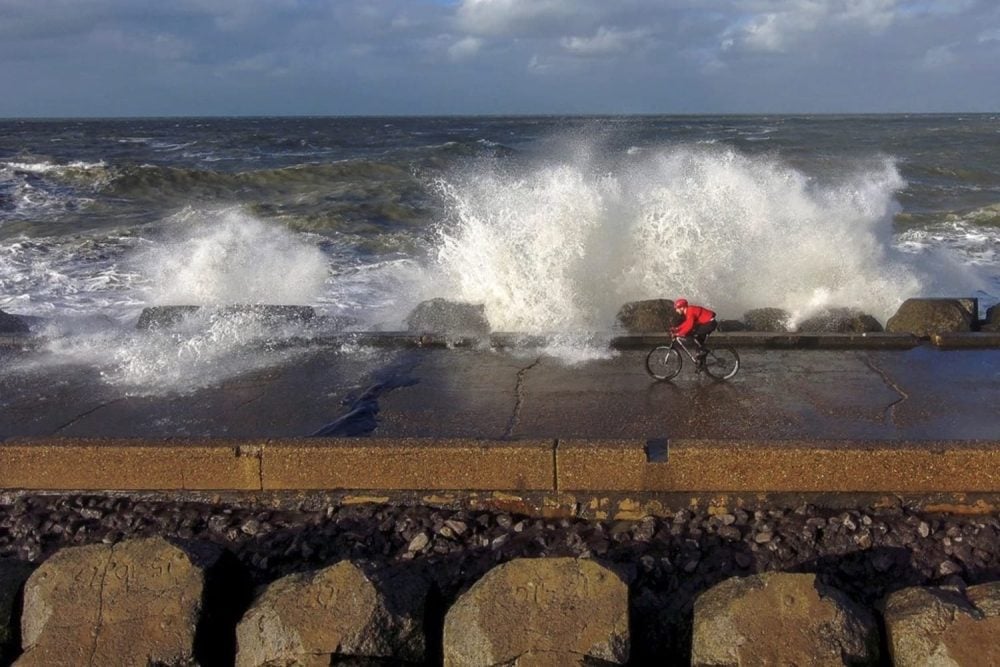 Cyclist in a red jacket riding a sea wall as waves burst over the barrier.