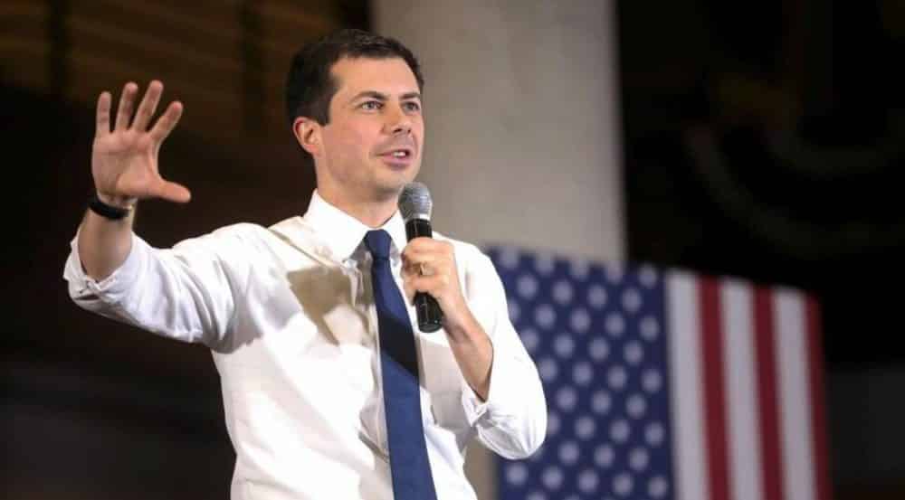 Pete Buttigieg gestures with a microphone while speaking in front of a US flag.