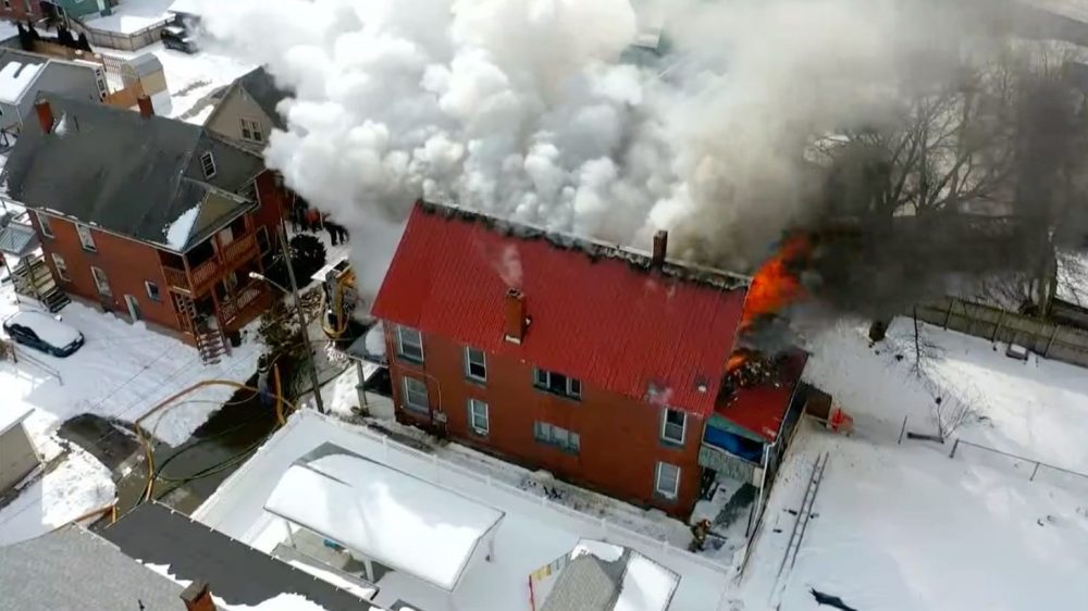 Aerial view of flames and smoke pouring from a red-roofed house in the snow.