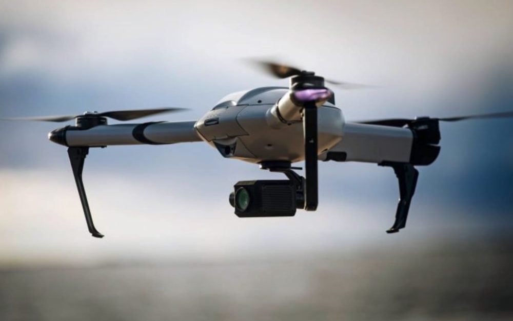 A silver quadcopter in flight carrying an oversized thermal imaging gimbal under its nose.