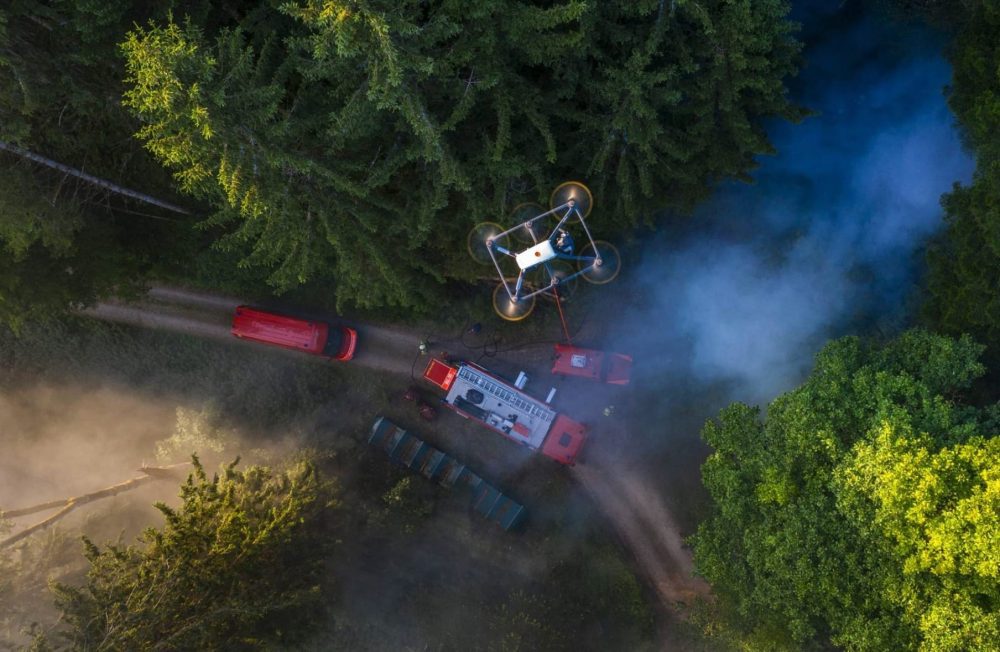 Overhead view of a tethered drone hovering above fire engines on a smoky forest road.