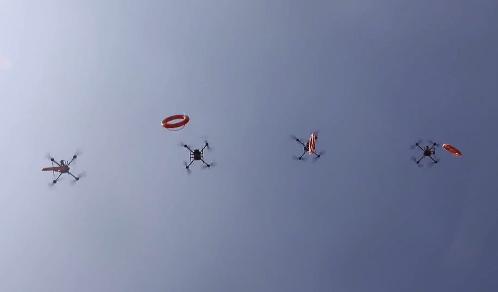 Four quadcopters flying in formation, each carrying an orange life ring beneath it.